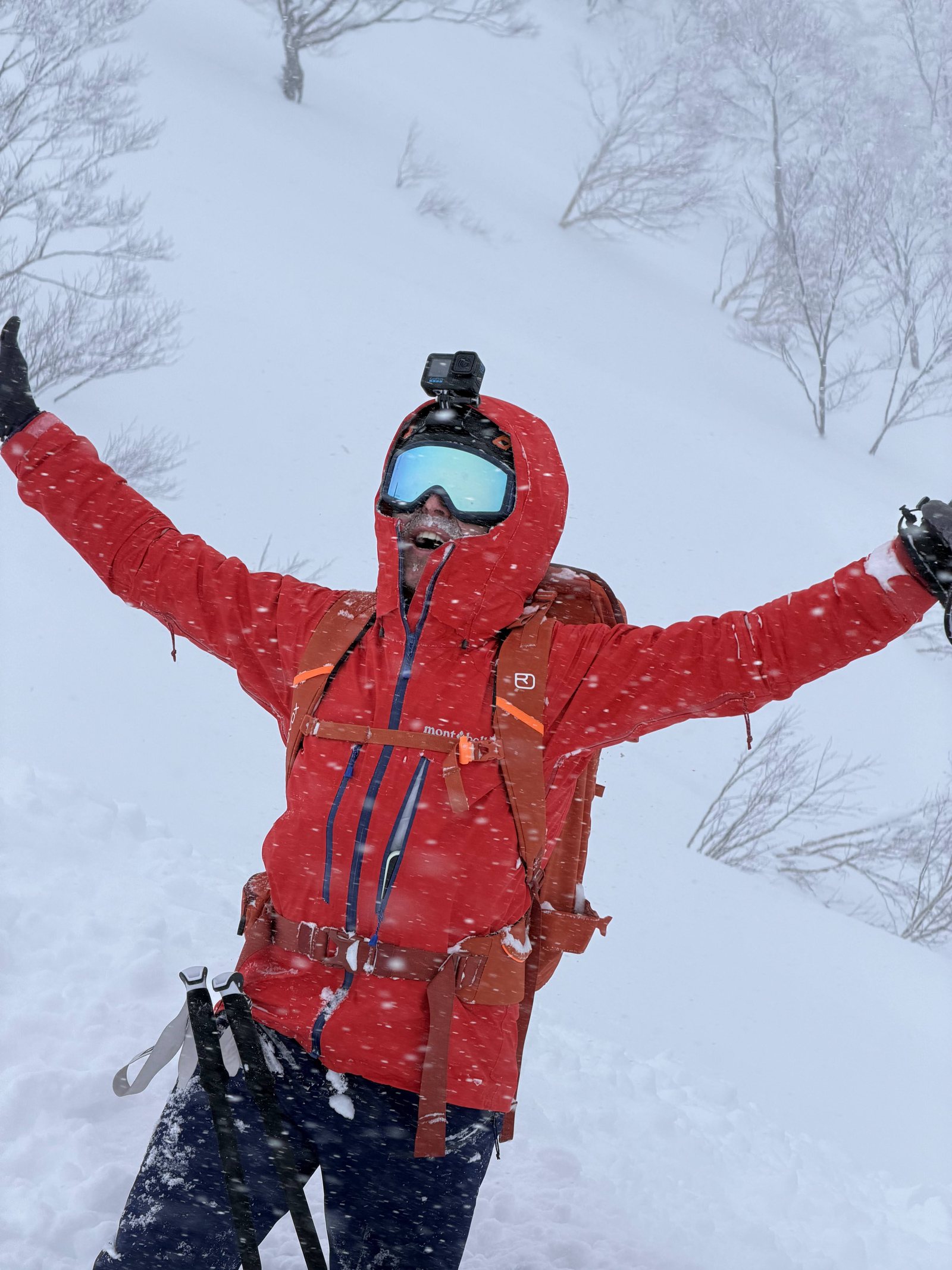 Sam in a red shell with arms wide open, laughing in heavy falling snow on a ski tour